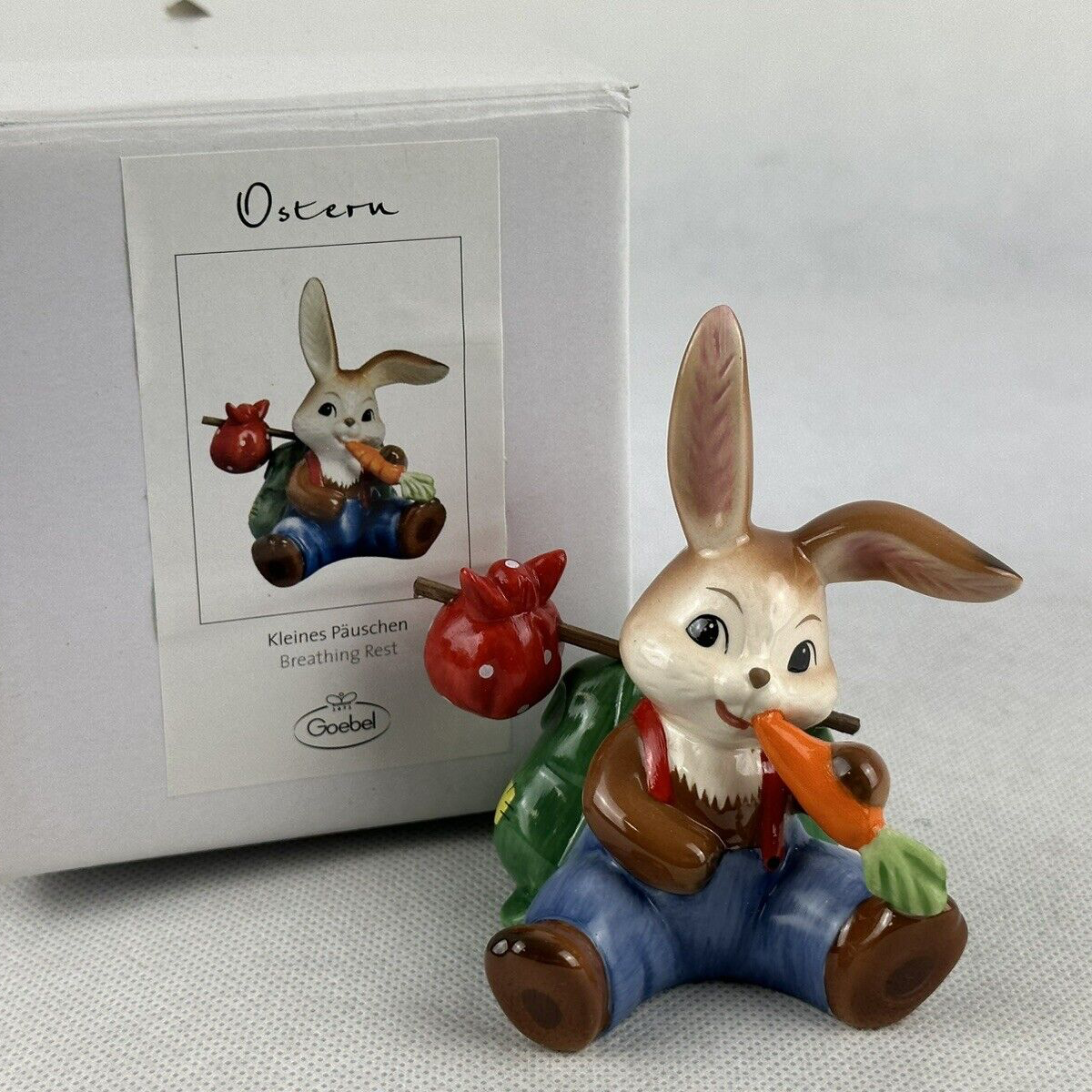 A porcelain figurine of a multicolored rabbit with a carrot, wearing blue overalls, sitting on a stool, positioned in front of its original box with a white background.